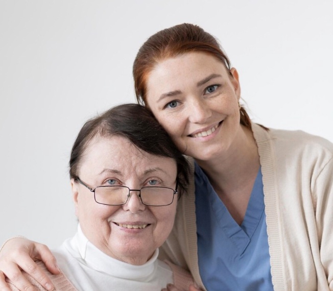 Cuidadora sorridente abraça uma senhora de óculos.