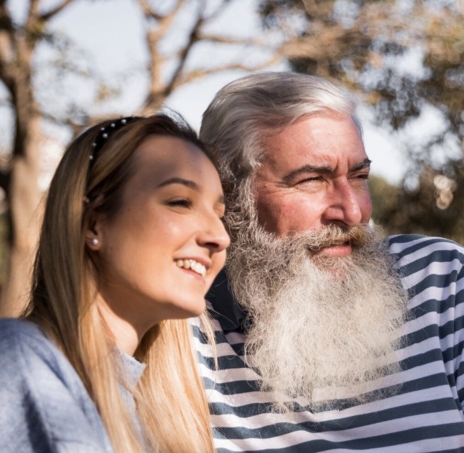 Filha adulta abraçada ao pai idoso, os dois sorrindo ao ar livre.